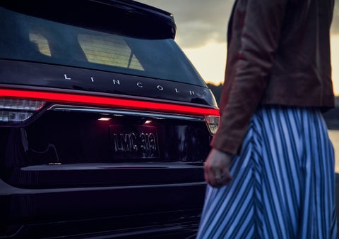 A person is shown near the rear of a 2024 Lincoln Aviator® SUV as the Lincoln Embrace illuminates the rear lights | West Point Lincoln of Sugar Land in Houston TX